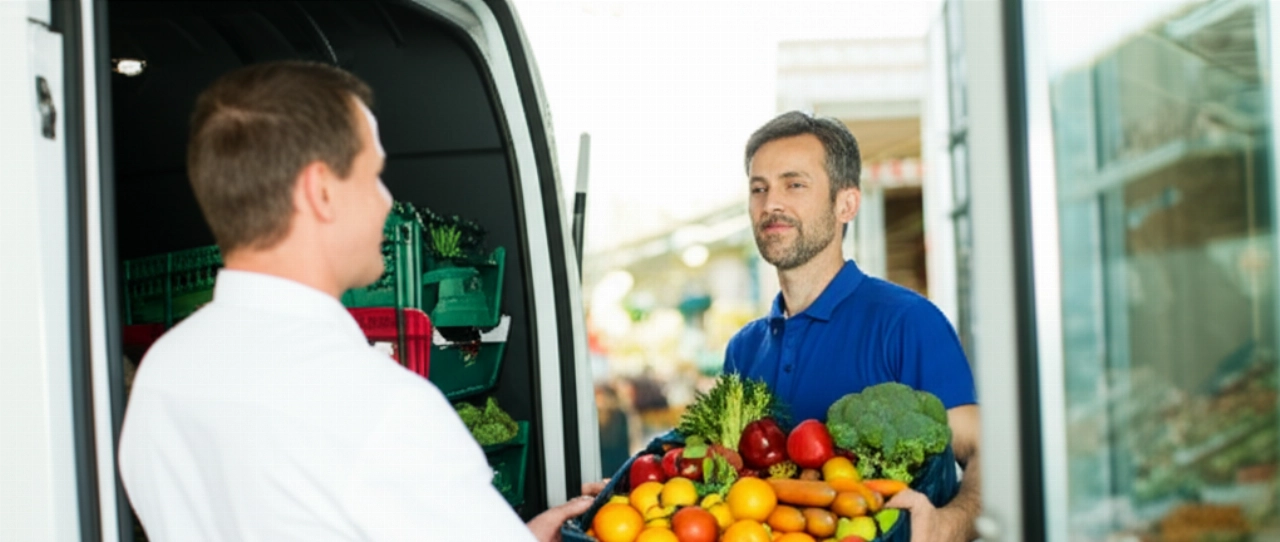 Autista caucasico che scarica frutta e verdura fresca da un furgone refrigerato, consegnandola a un cuoco o gestore di negozio, illustrando la fase finale del trasporto alimentare e la freschezza dei prodotti.